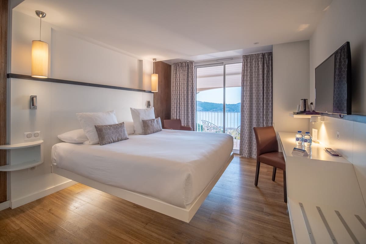 A modern hotel room with a large bed, a desk with a chair, a flat-screen TV, and a view of the outdoors.