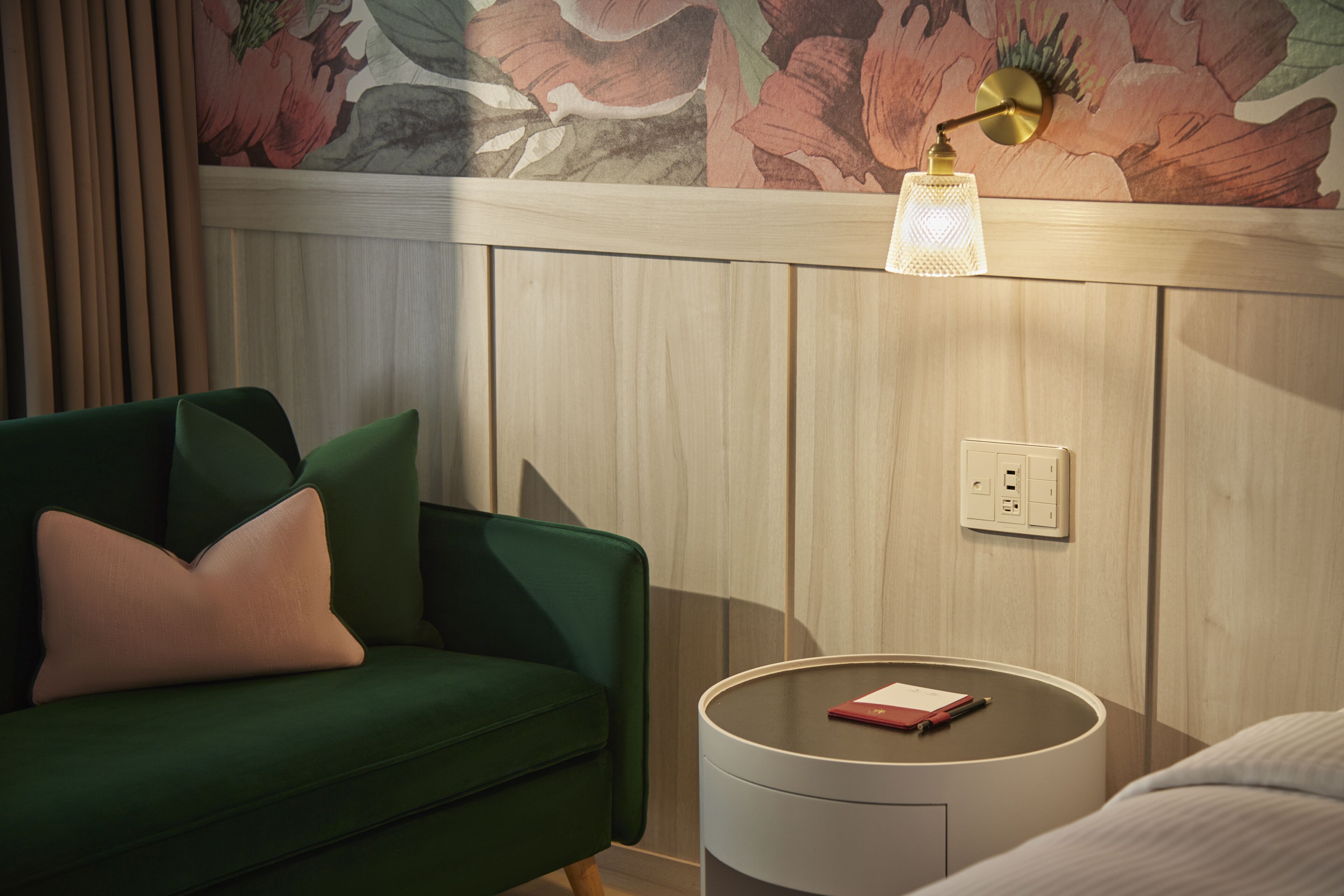 Un sofá verde con una sola almohada, una mesita redonda con una lámpara y papel tapiz floral sobre un panel de madera.