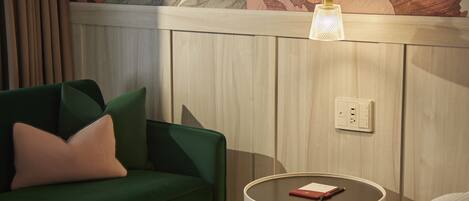 A green sofa with a single pillow, a round side table with a lamp, and a floral wallpaper above a wooden panel wall.