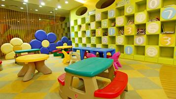 Zona de juegos infantiles con mesas y sillas de colores, una pared con estantes numerados y un suelo a cuadros.
