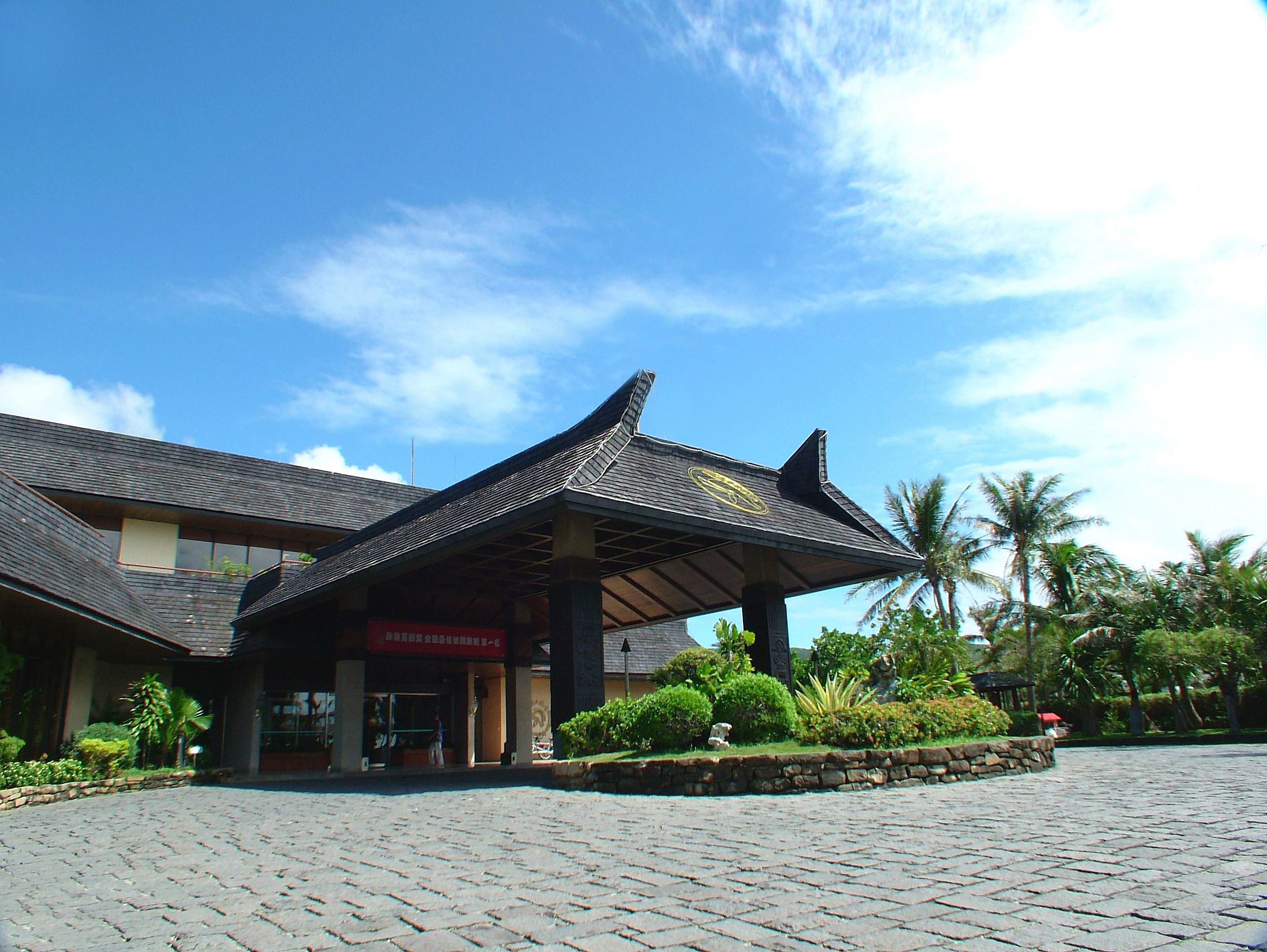 A building with a traditional roof structure, a paved entrance area, and surrounding greenery.