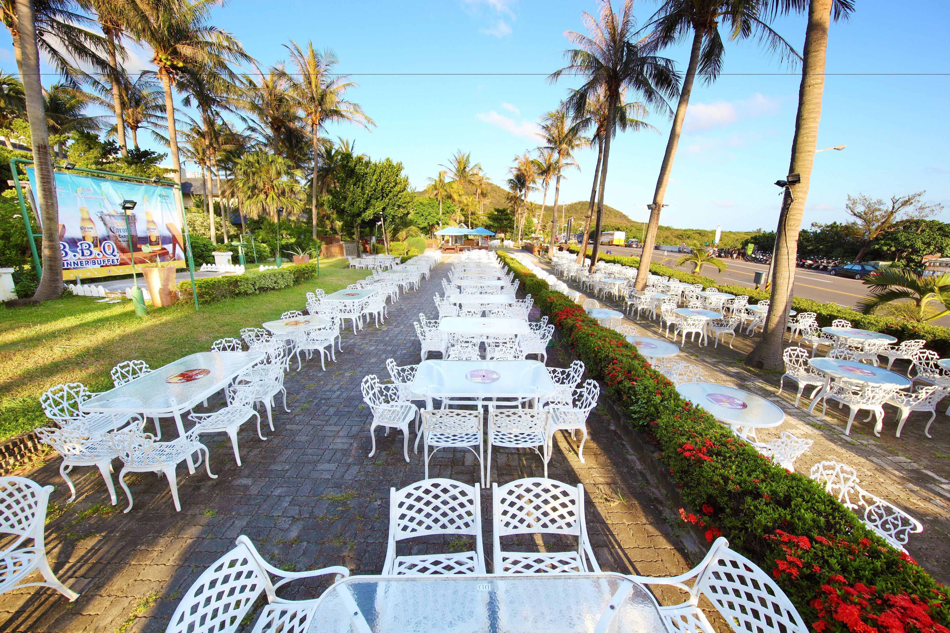 An outdoor dining area with white metal tables and chairs arranged in rows, surrounded by palm trees and greenery.