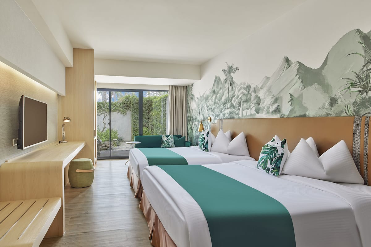 A hotel room with two beds, a TV, a desk, and a large window with a view of greenery.