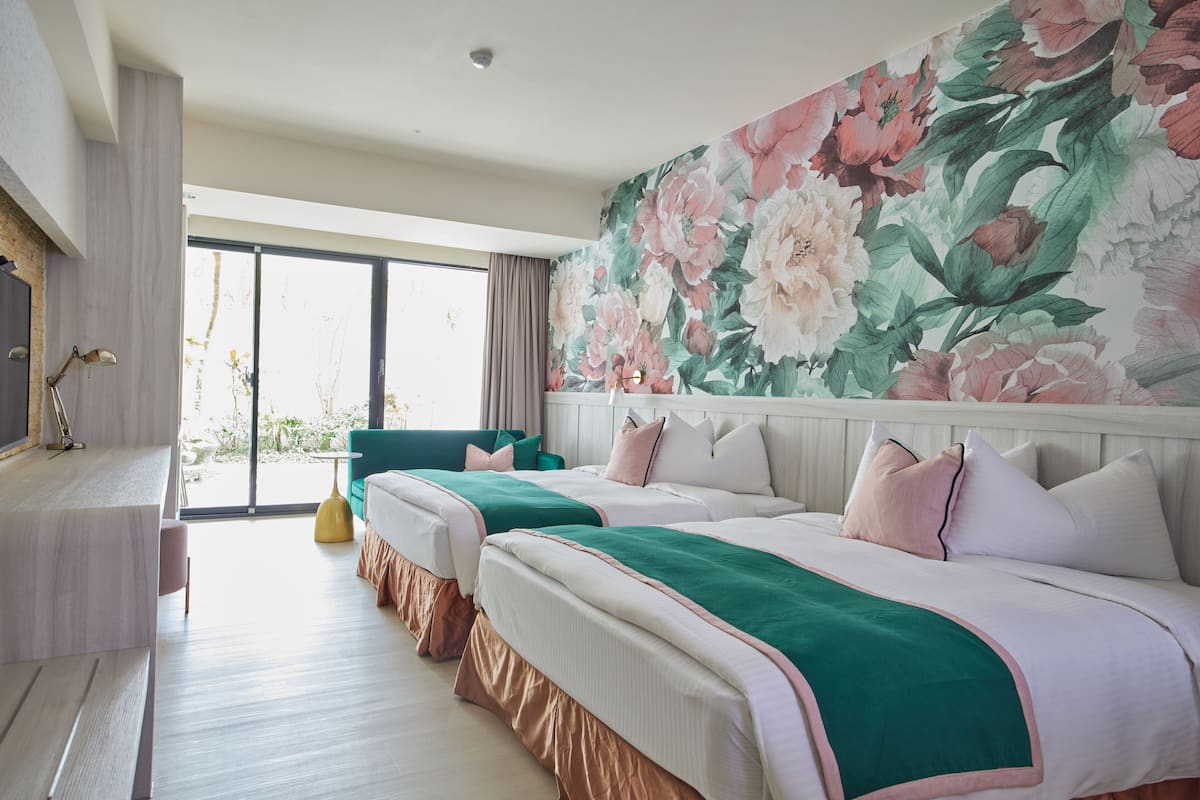 A hotel room with two beds, a large floral wall mural, a sliding glass door to the outside, and a small table with a lamp.