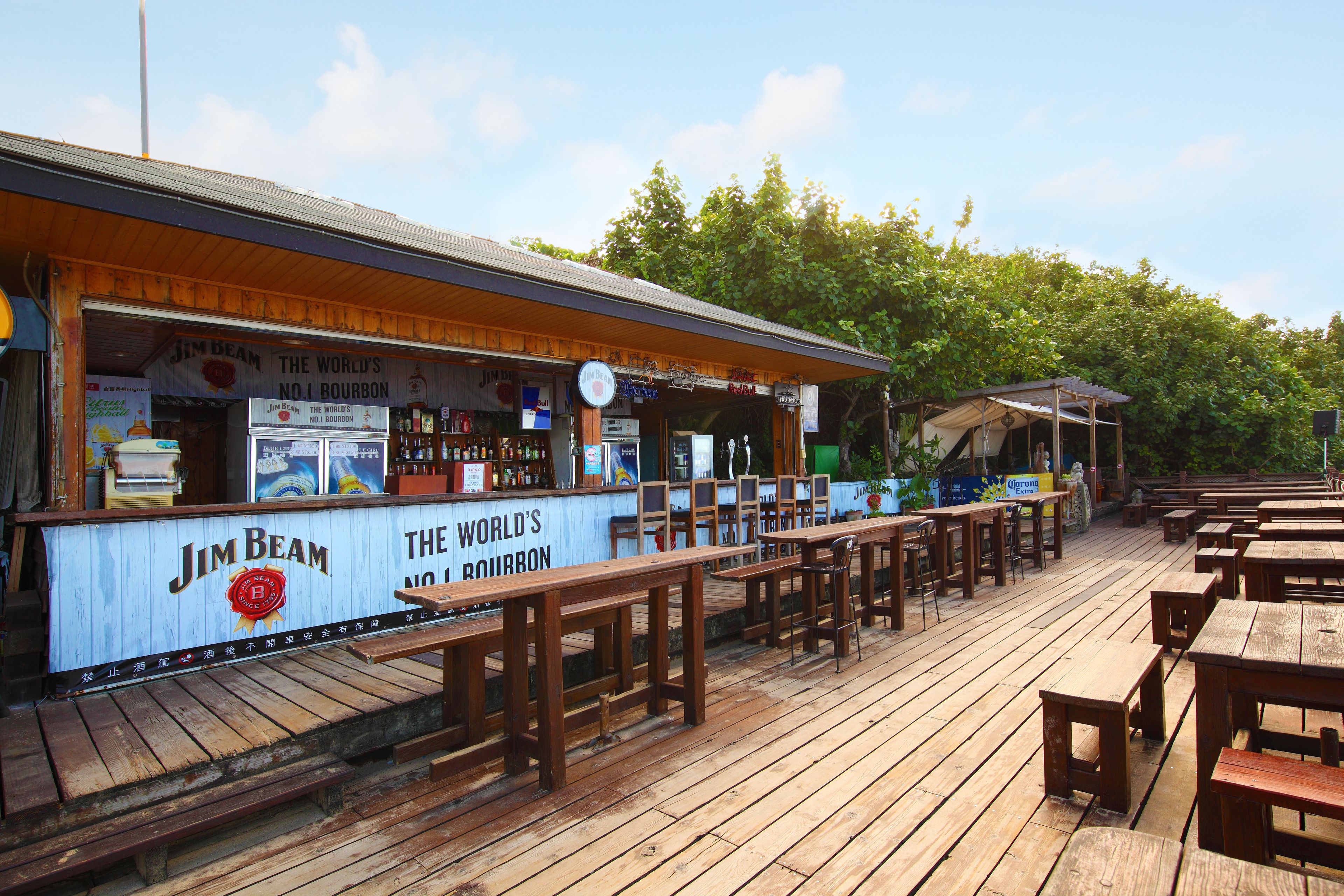 Uma área externa com mesas e bancos de madeira, uma seção coberta e uma placa da Jim Beam.