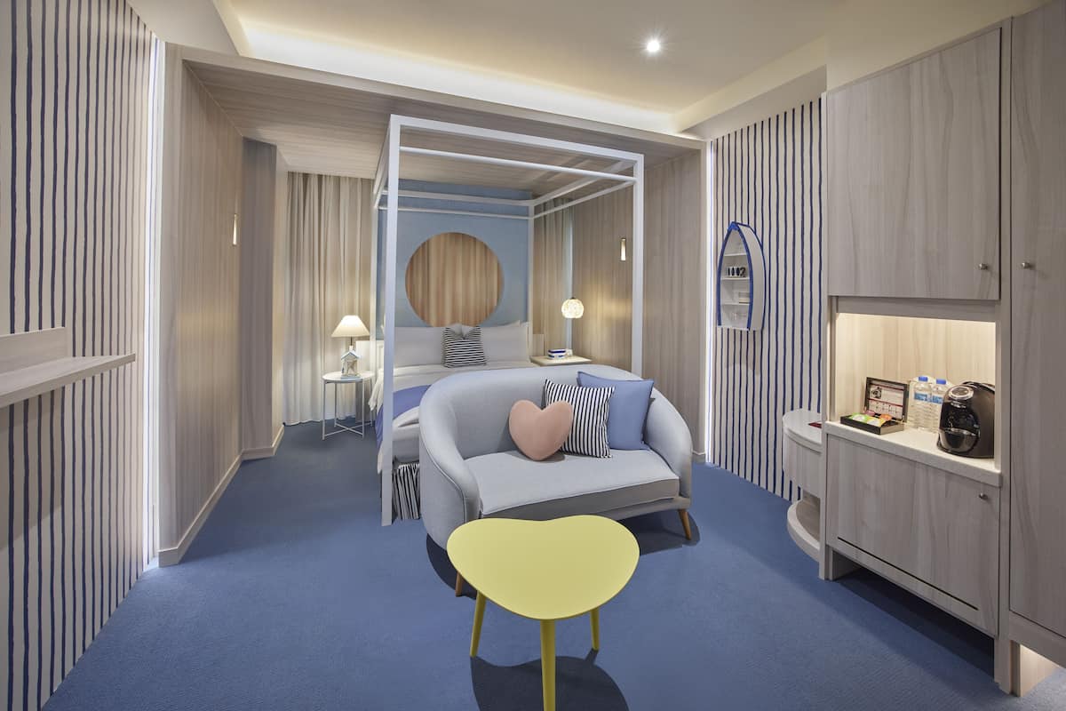 A modern hotel room with a blue carpet, a bed with a heart-shaped pillow, a yellow chair, a bedside table with a lamp, and a wardrobe.