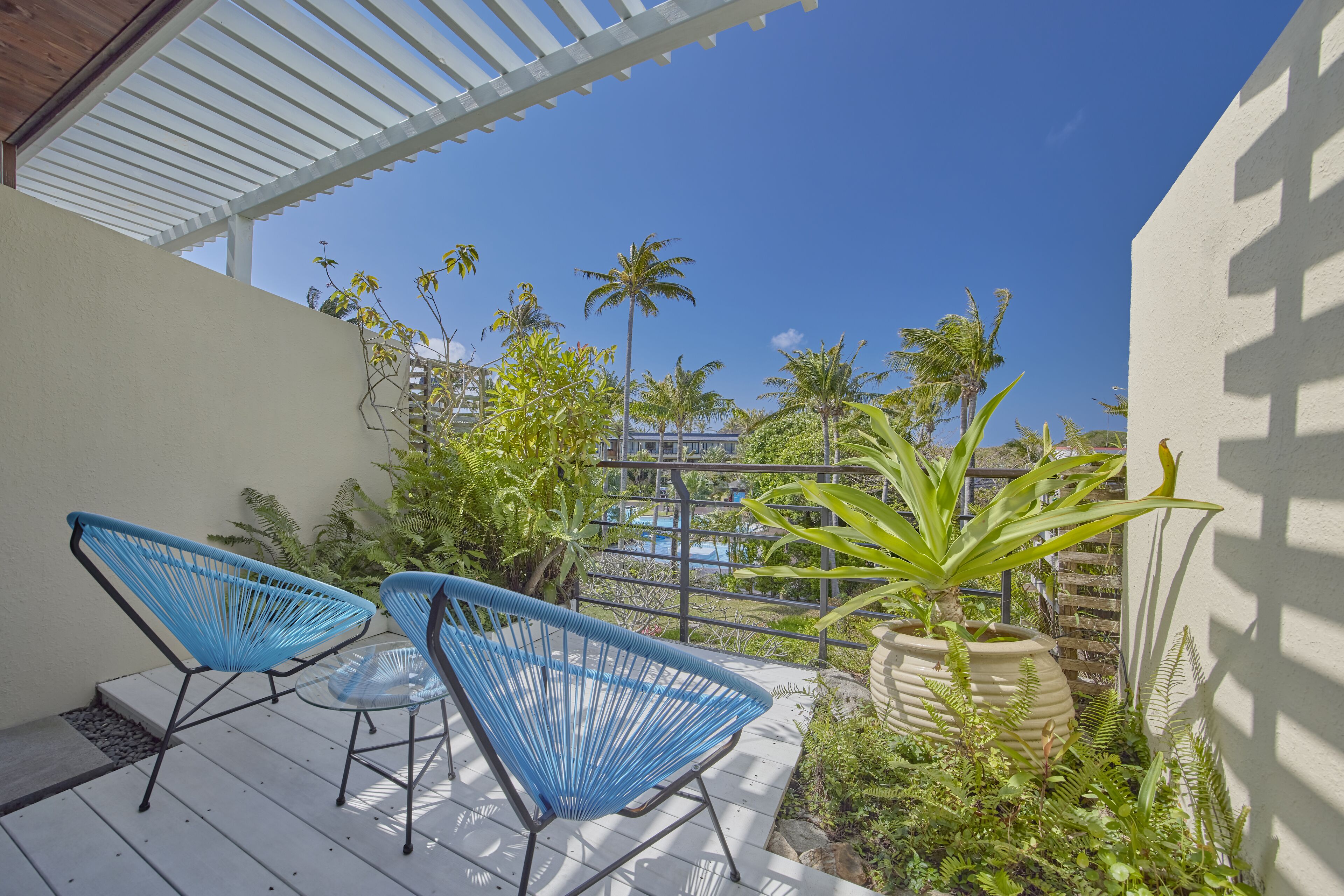 A patio with two blue chairs and a table, surrounded by greenery and a view of a pool.