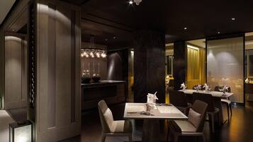 Un comedor moderno con una mesa central dispuesta para dos personas, rodeada de sillas. Hay lámparas colgantes sobre la mesa y una zona de bar con taburetes a la izquierda. La habitación tiene paneles de madera oscura y una partición de vidrio a la derecha.