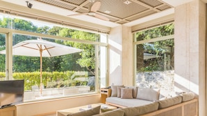 Una sala de estar moderna con un sofá grande, una mesa de centro y un televisor. Amplias ventanas ofrecen vistas a zonas verdes exteriores.