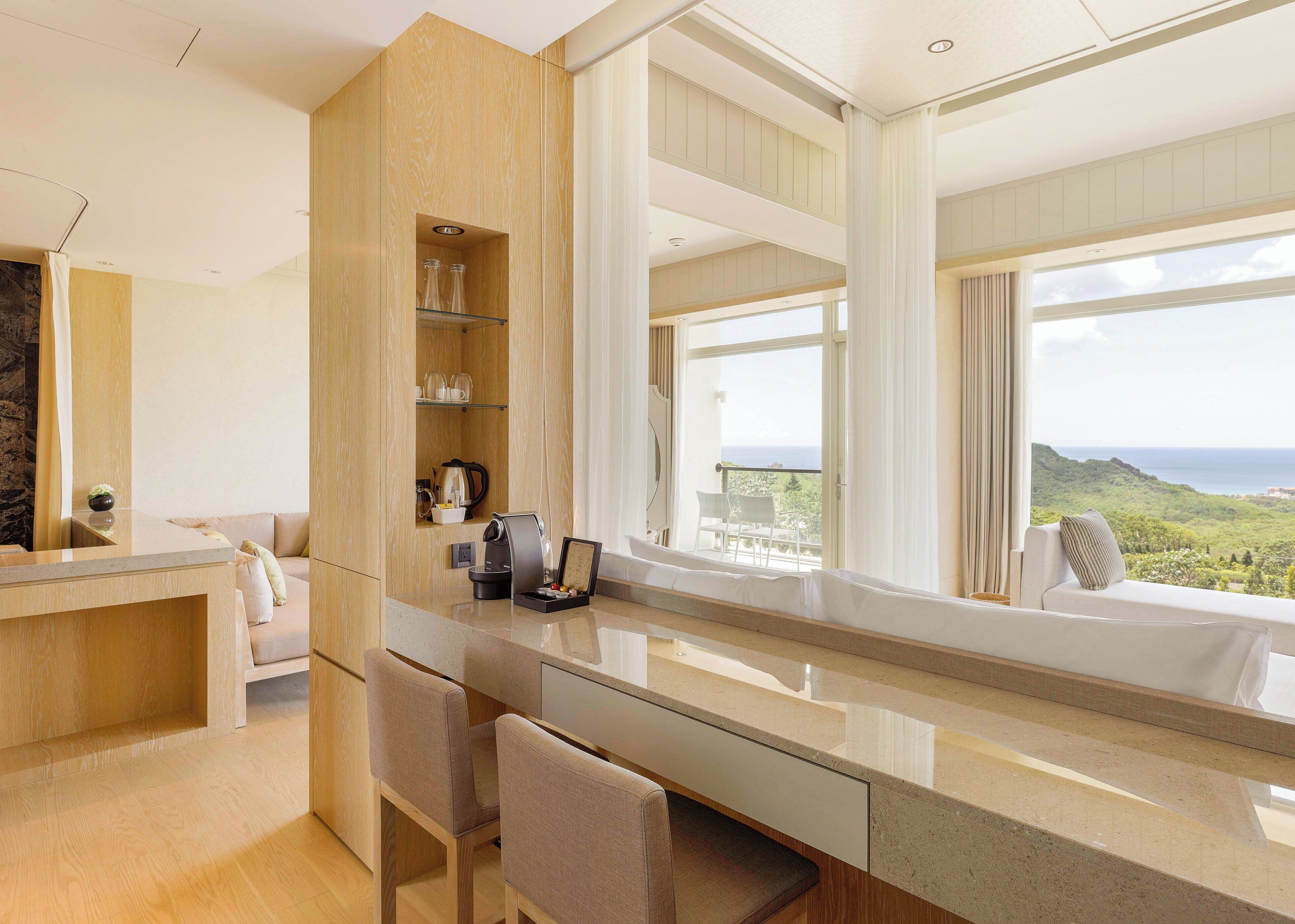 Una habitación de hotel moderna con un gran ventanal que ofrece vistas panorámicas, un mostrador de recepción de madera y una zona de estar con máquina de café.