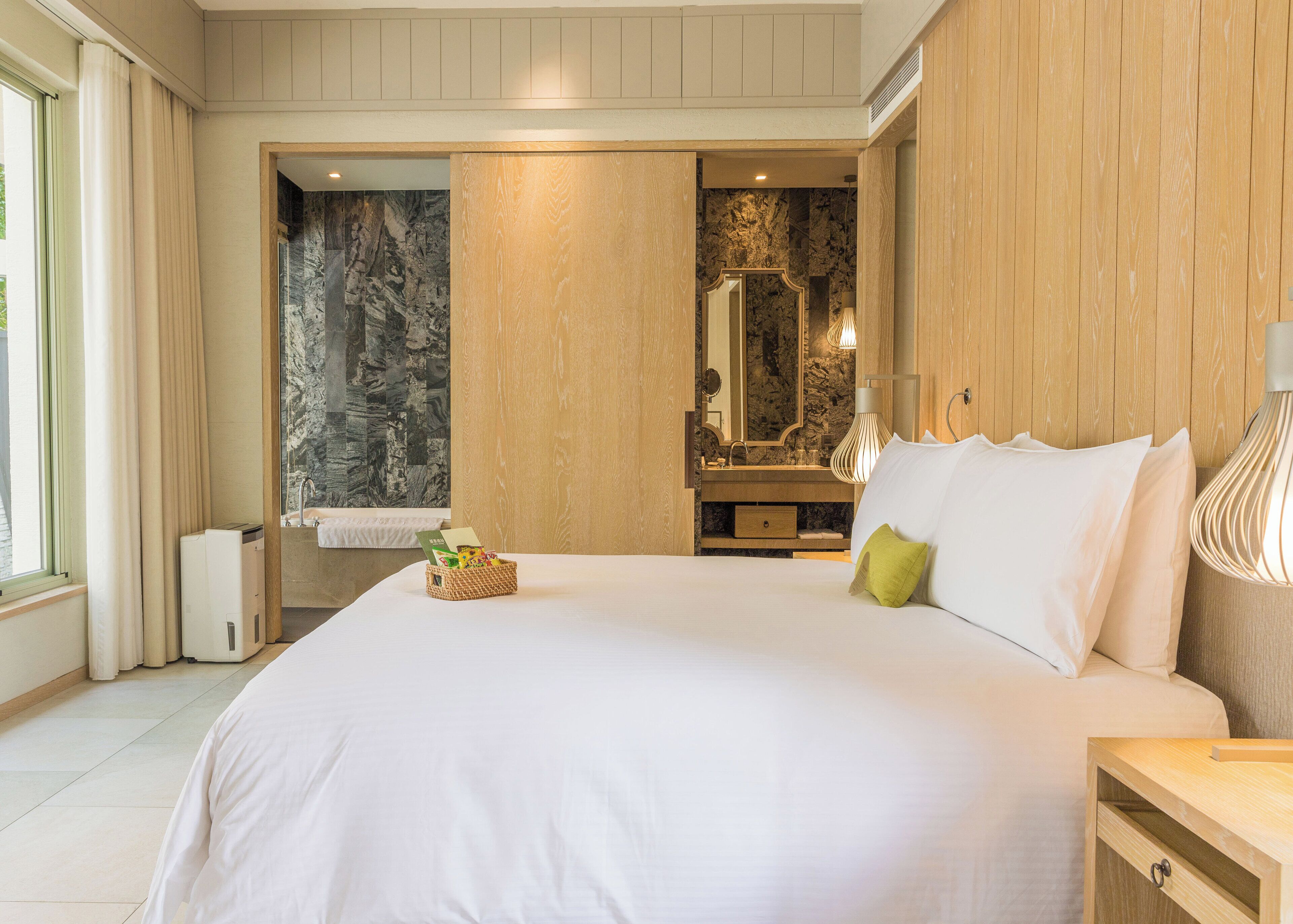 A bedroom with a large bed, wooden paneling, a marble wall, a bathroom with a sink and mirror, and a bedside table with a lamp.