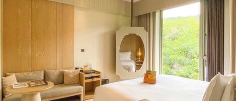 A hotel room with a bed, a sofa, a small table, a mirror, and a window with a view of greenery.