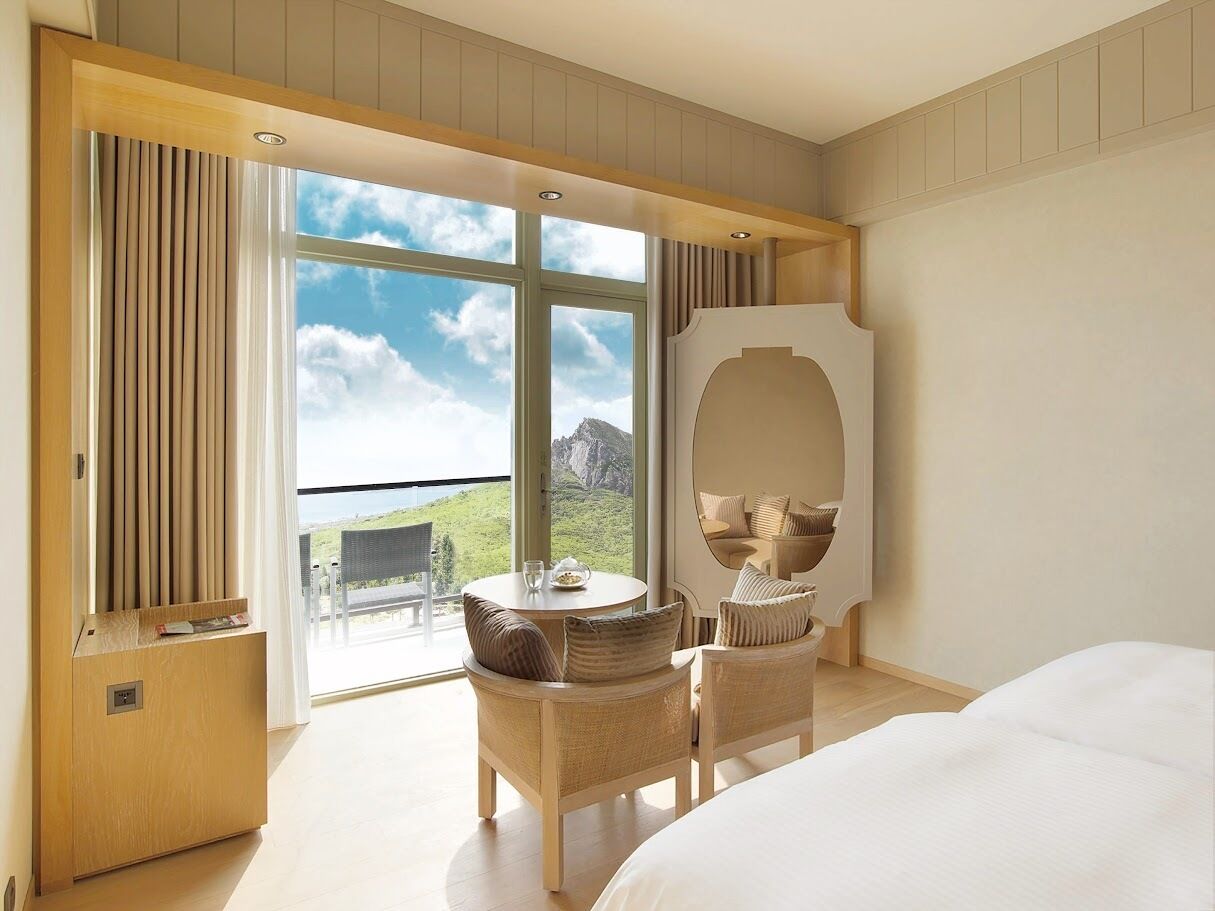 Habitación de hotel con una cama grande, una mesa redonda con sillas, un espejo y vistas a un paisaje montañoso a través de ventanas amplias.