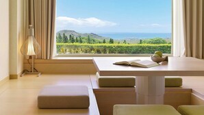 Una habitación con un ventanal grande que ofrece vistas a un paisaje verde y montañas. Hay una mesa con un libro abierto y un cuenco encima.