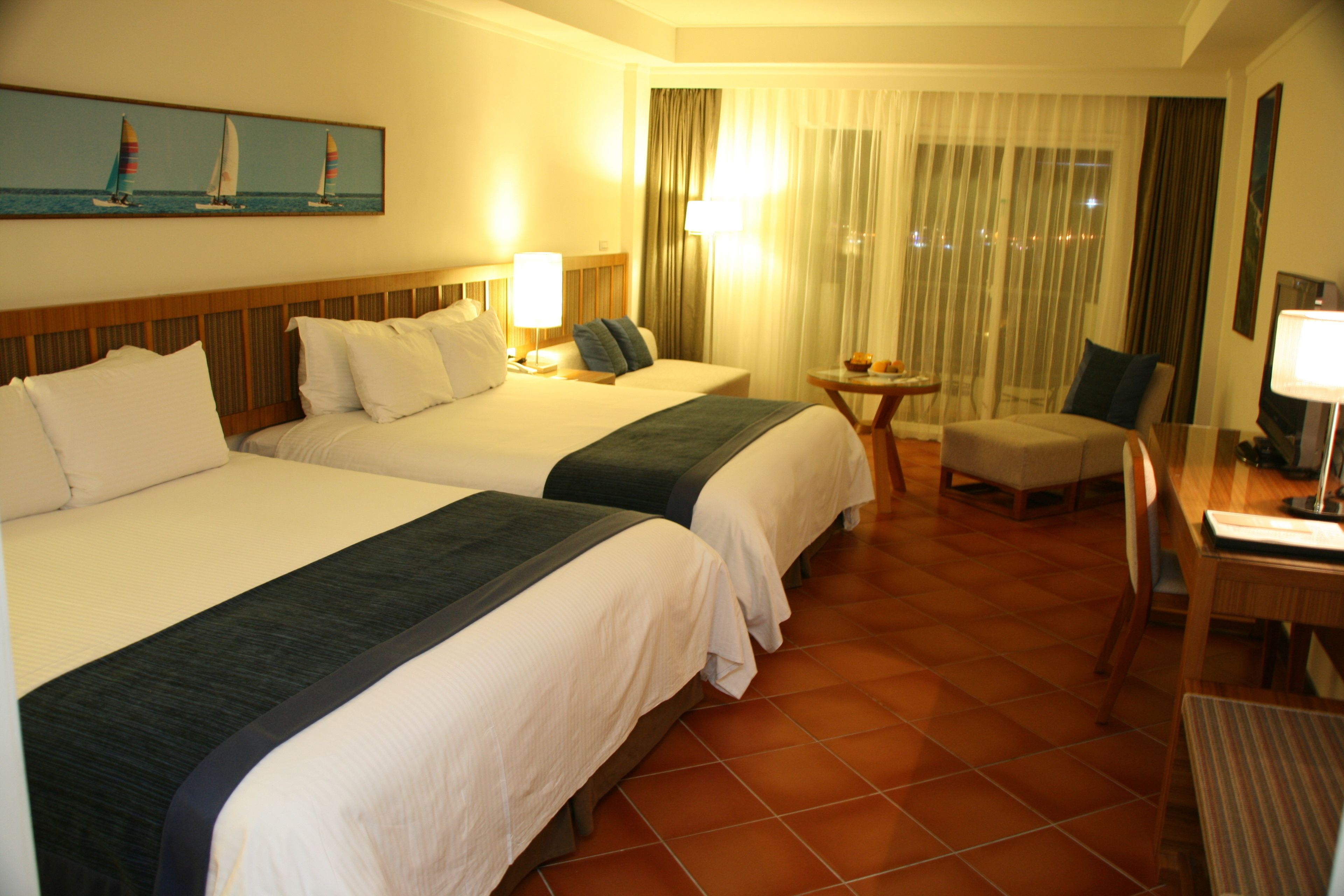 A hotel room with two beds, a small table, a chair, and a desk with a lamp.