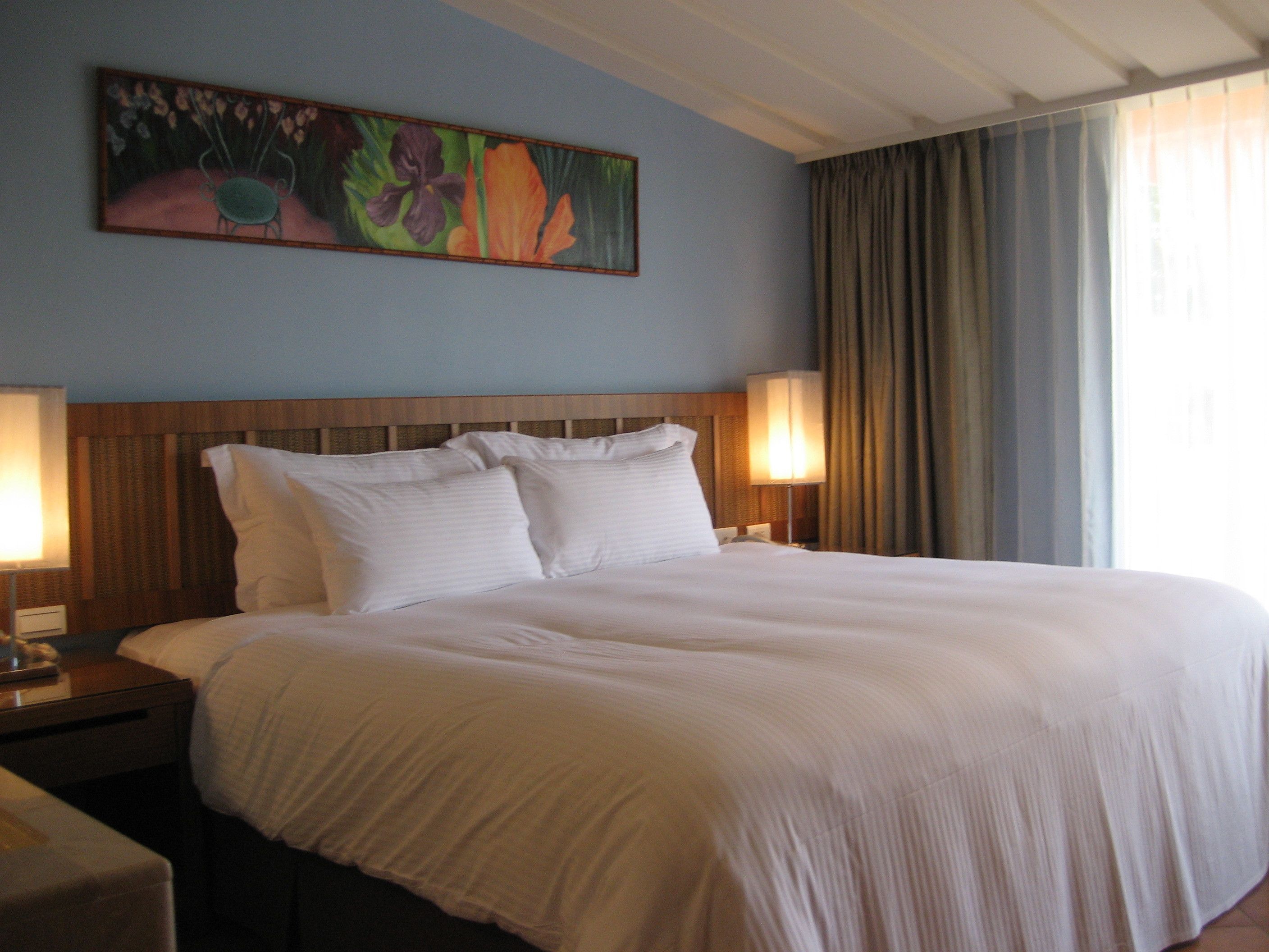 A hotel room with a large bed, two bedside lamps, a wooden headboard, and a floral painting on the wall.