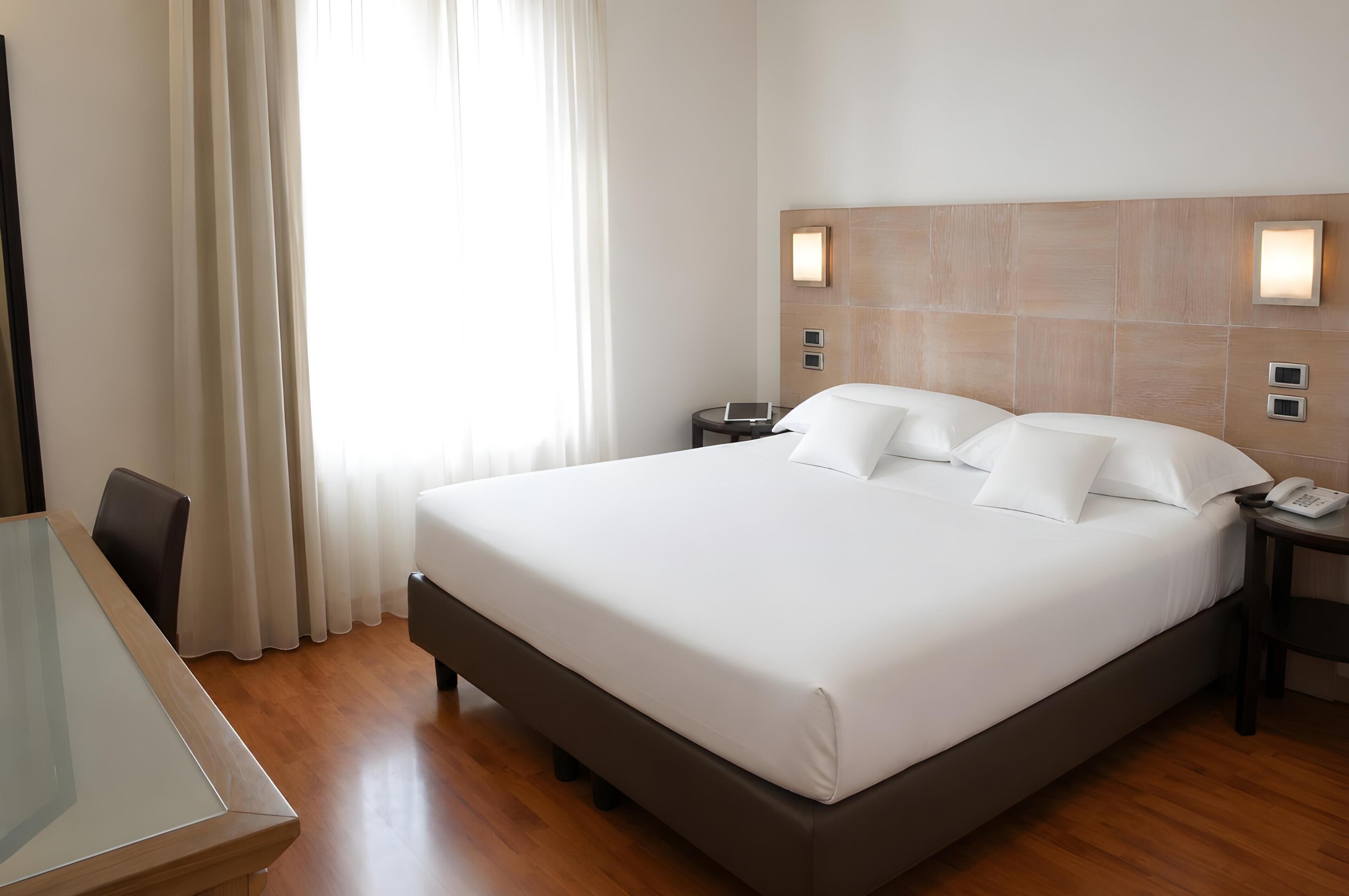 Quarto de hotel com uma cama grande, uma mesa de cabeceira com telefone, uma pequena mesa com cadeira e uma janela com cortinas.