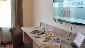 A hotel room with a desk, a television, a remote control, a tablet, a cup, a plate with pastries, a chair, and a window with curtains.