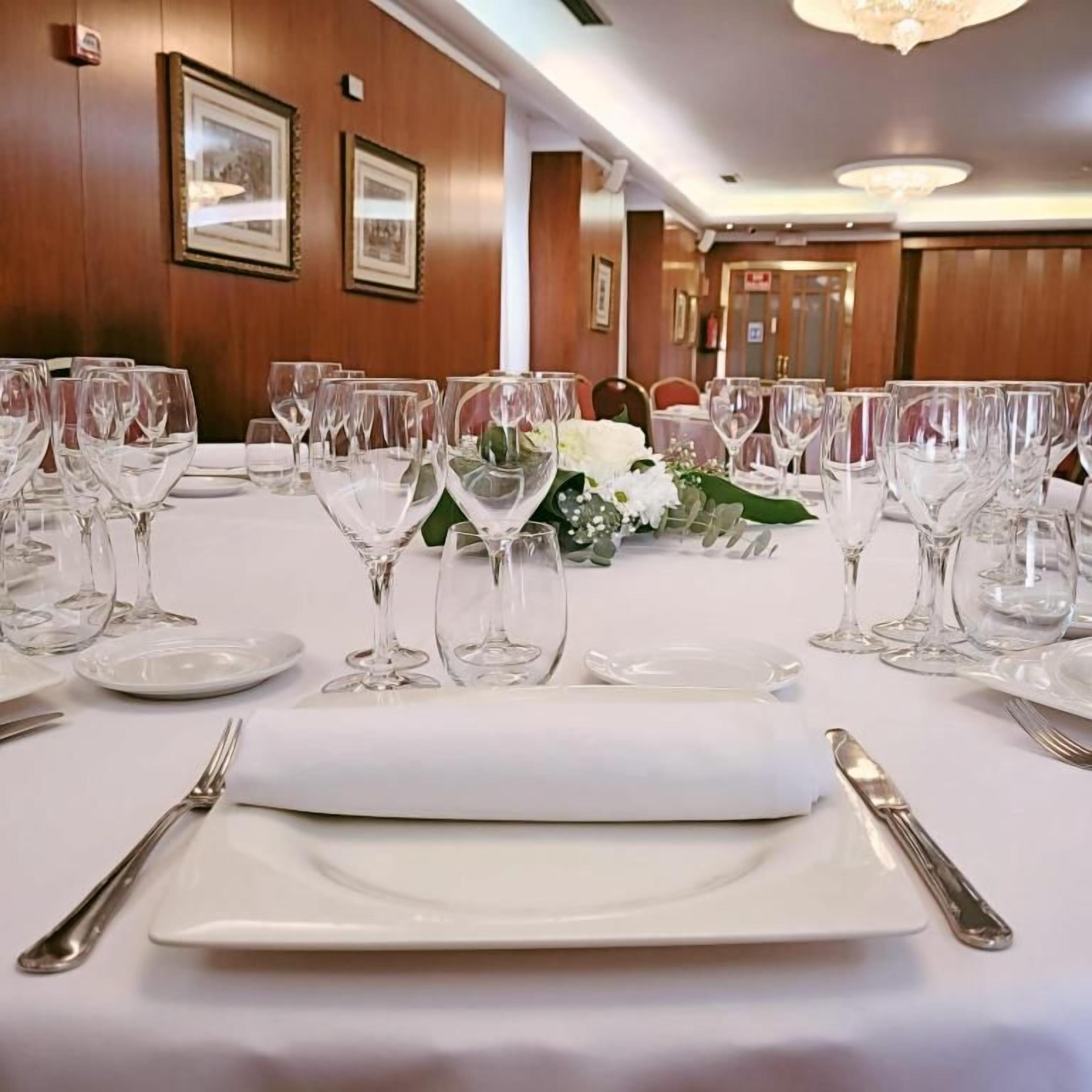 Una mesa de comedor dispuesta con platos blancos, cubiertos y copas de vino, con un mantel blanco y un pequeño arreglo floral.