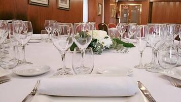 Una mesa de comedor dispuesta con platos blancos, cubiertos y copas de vino, con un mantel blanco y un pequeño arreglo floral.