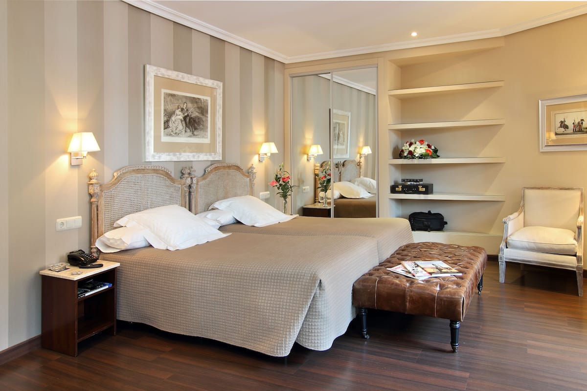 Una habitación de hotel ordenada con una cama grande, mesitas de noche, una silla, un banco y un espejo.