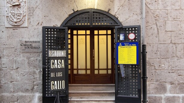The entrance to Casa de los Gallo, a restaurant with a traditional facade, a coat of arms above the door, and a menu sign on the right. - Hotel Imperial (Valladolid)