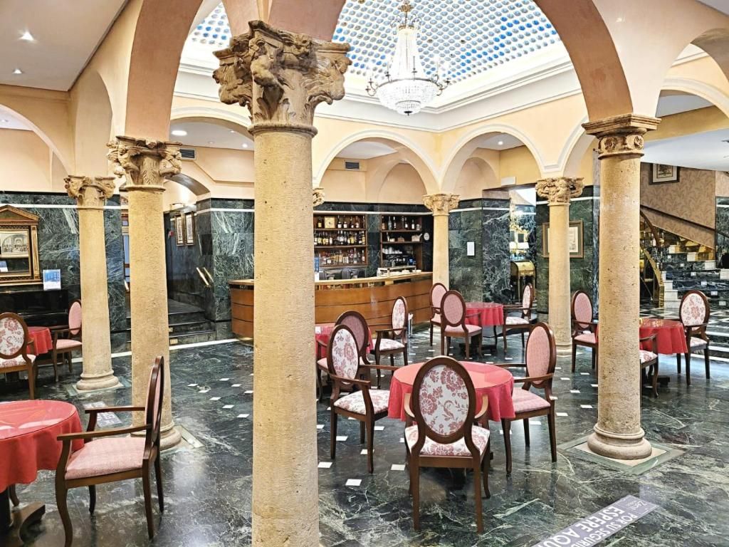 Un vestíbulo amplio con piso de mármol, mesas rojas y sillas clásicas, que presenta una columna central y un candelabro.