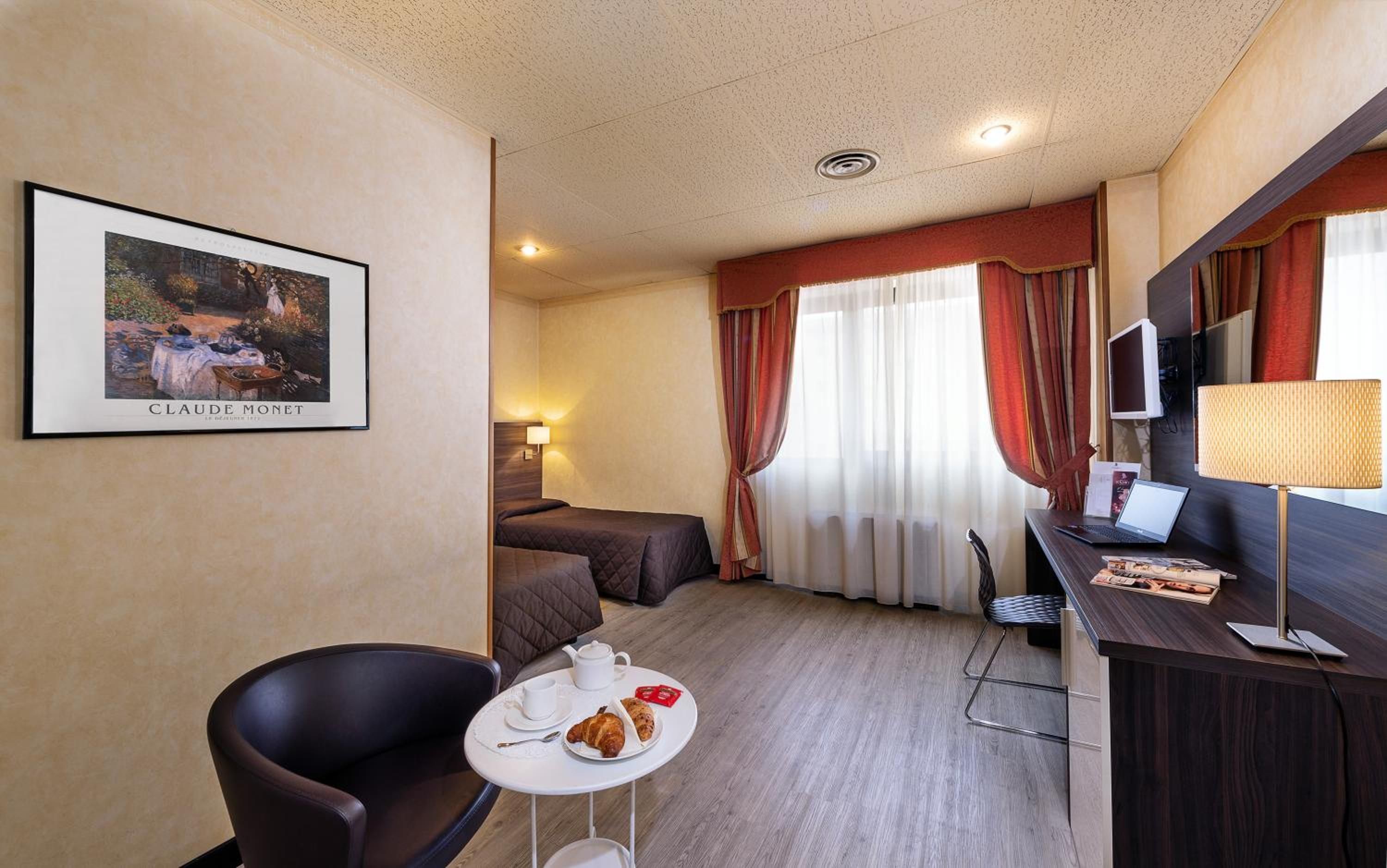 Quarto de hotel com uma cama, escrivaninha com um laptop, cadeira, pequena mesa com uma chávena e pastelaria, e uma fotografia emoldurada na parede.