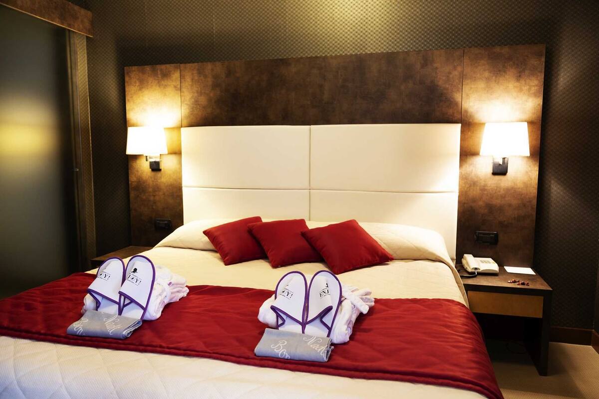 Habitación de hotel con una cama grande, ropa de cama roja y blanca, dos almohadas rojas y dos toallas con el logo 'NAT'. Hay dos mesitas de noche con lámparas y un teléfono.