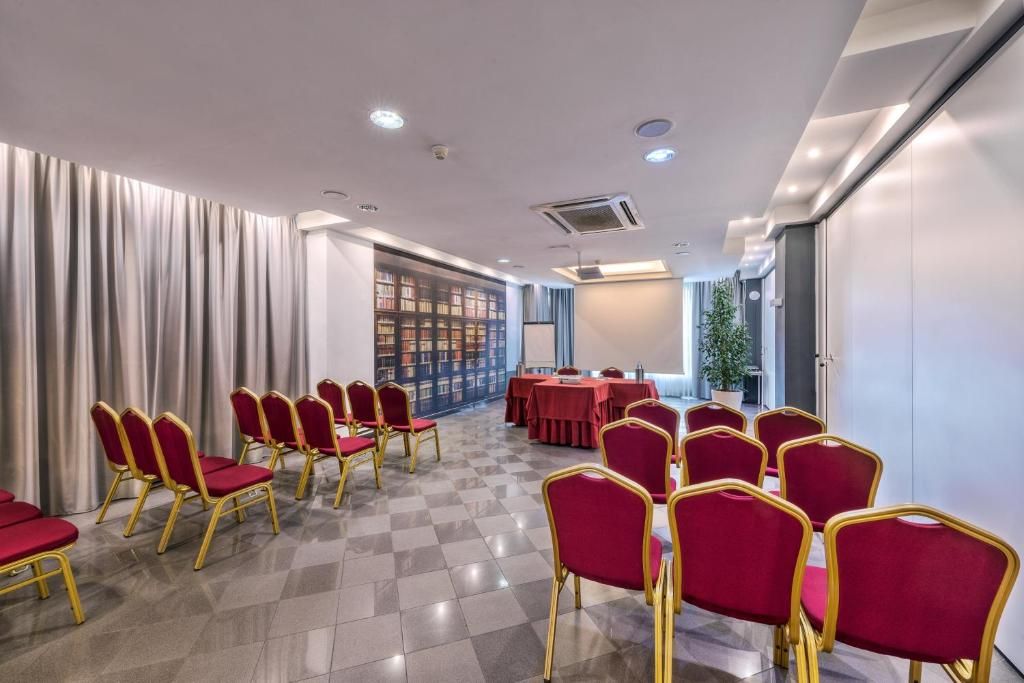 A conference room with rows of red chairs, a long table, and a bookshelf in the background.