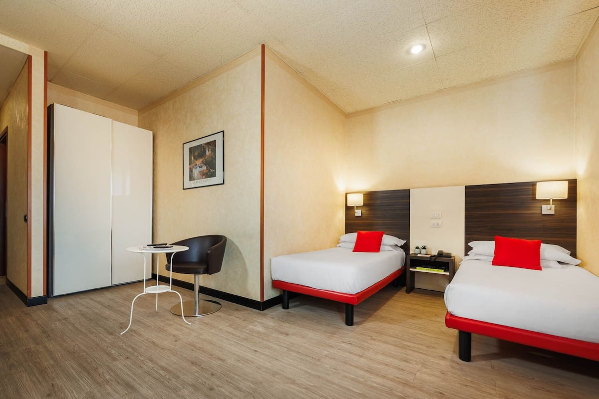 Habitación de hotel con dos camas, una mesita, una silla y un armario.