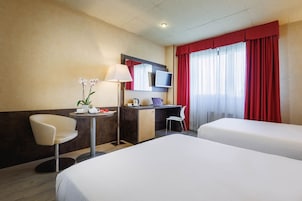 Quarto de hotel com duas camas, uma escrivaninha, uma cadeira, um abajur e uma televisão.