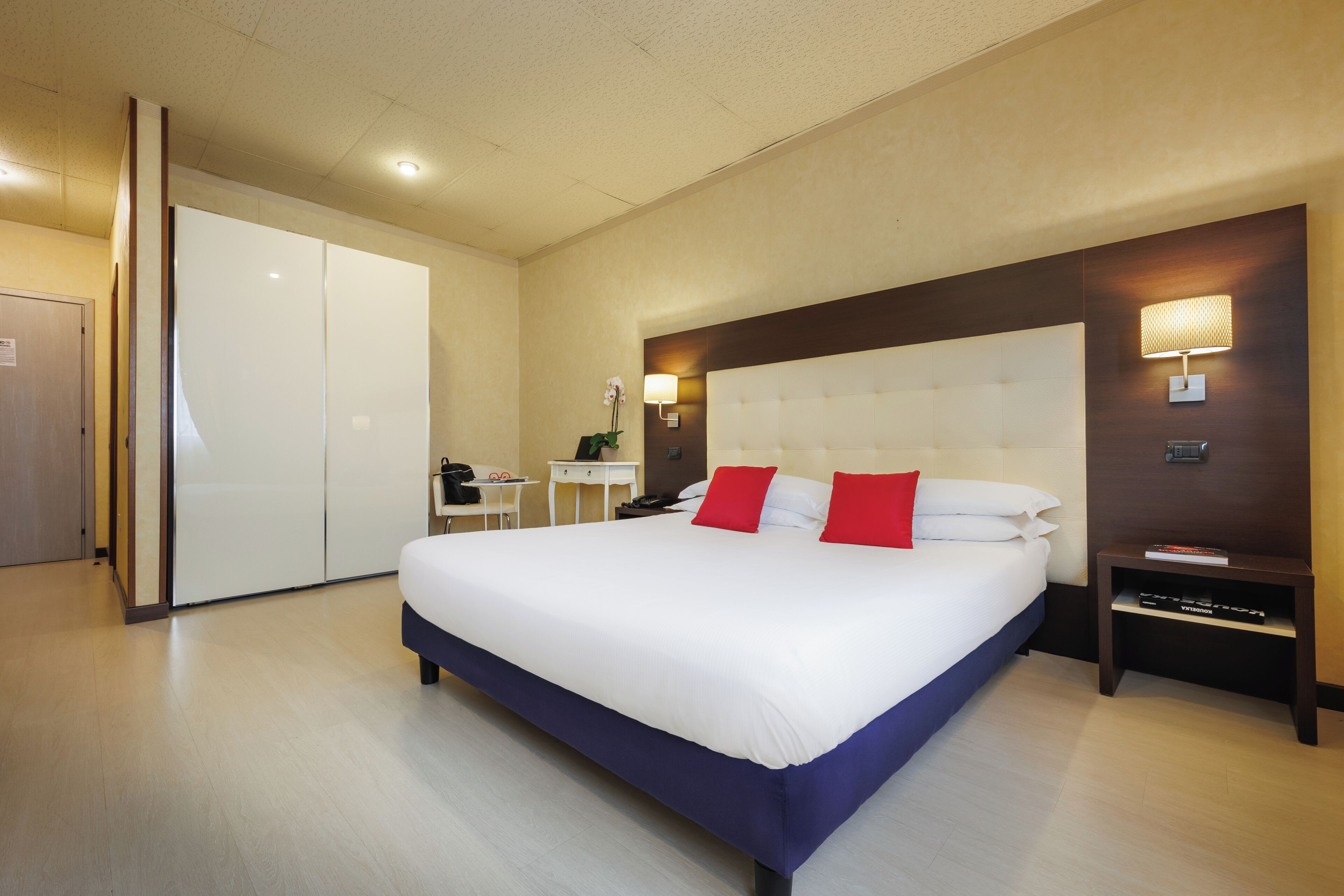 A hotel room with a large bed, a desk, a chair, a wardrobe, and a nightstand with a lamp.
