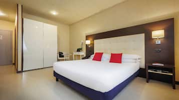 A hotel room with a large bed, a desk, a chair, a wardrobe, and a nightstand with a lamp.