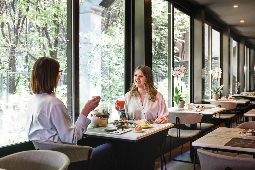 Een café met tafels en stoelen, grote ramen en uitzicht op groen.
