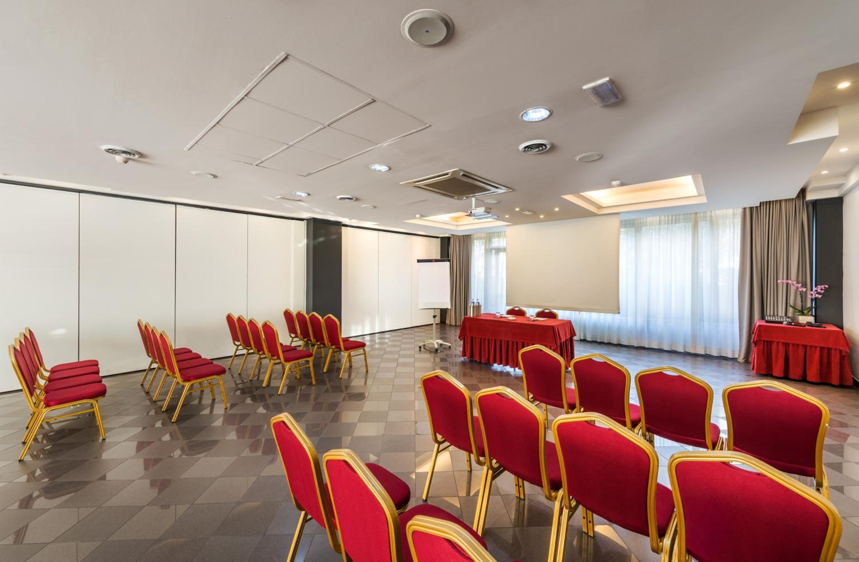 Uma sala de conferências com fileiras de cadeiras vermelhas, uma mesa com toalha vermelha e uma tela de projetor.