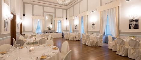 A banquet hall with round tables, white chairs, chandeliers, and framed pictures on the walls.