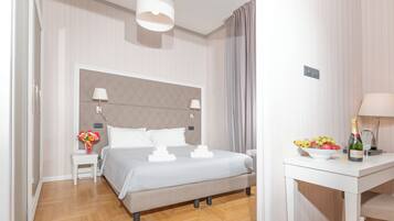 Une chambre d’hôtel moderne avec un grand lit, une table de chevet avec une lampe, une petite table avec un panier de fruits et une bouteille, et un rideau à la fenêtre.