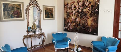 Una habitación con tres sillones azules, una mesa de centro de cristal, un cuadro floral grande y dos obras de arte enmarcadas en la pared.