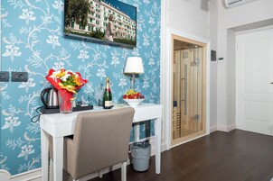 Une chambre d’hôtel avec une table blanche, un bouquet de fleurs, une bouteille de champagne et un panier de fruits.