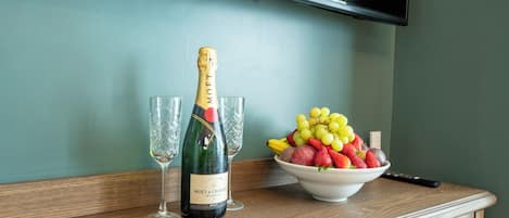 Une bouteille de champagne Moët & Chandon, deux verres et un saladier de fruits sur une table en bois, le tout placé devant un mur de couleur turquoise, avec un téléviseur au-dessus.