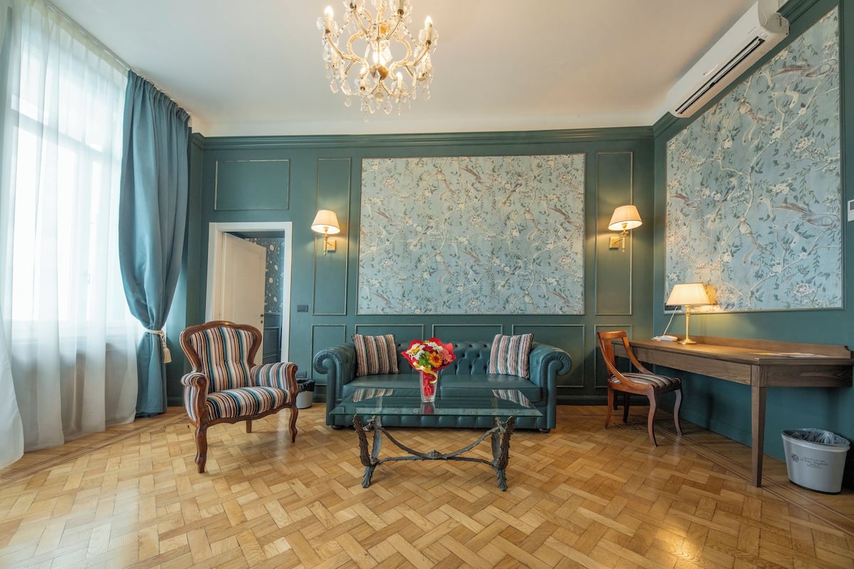 Ein Zimmer mit Fischgrätparkett, einem grünen Sofa mit gestreiften Kissen, einem Holztisch und einer Pendelleuchte.