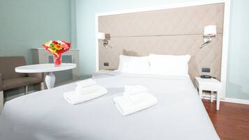 Ein Hotelzimmer mit einem großen Bett, zwei weißen Nachttischen, einem kleinen runden Tisch mit Blumenvase, einer Couch und einer Wandlampe.