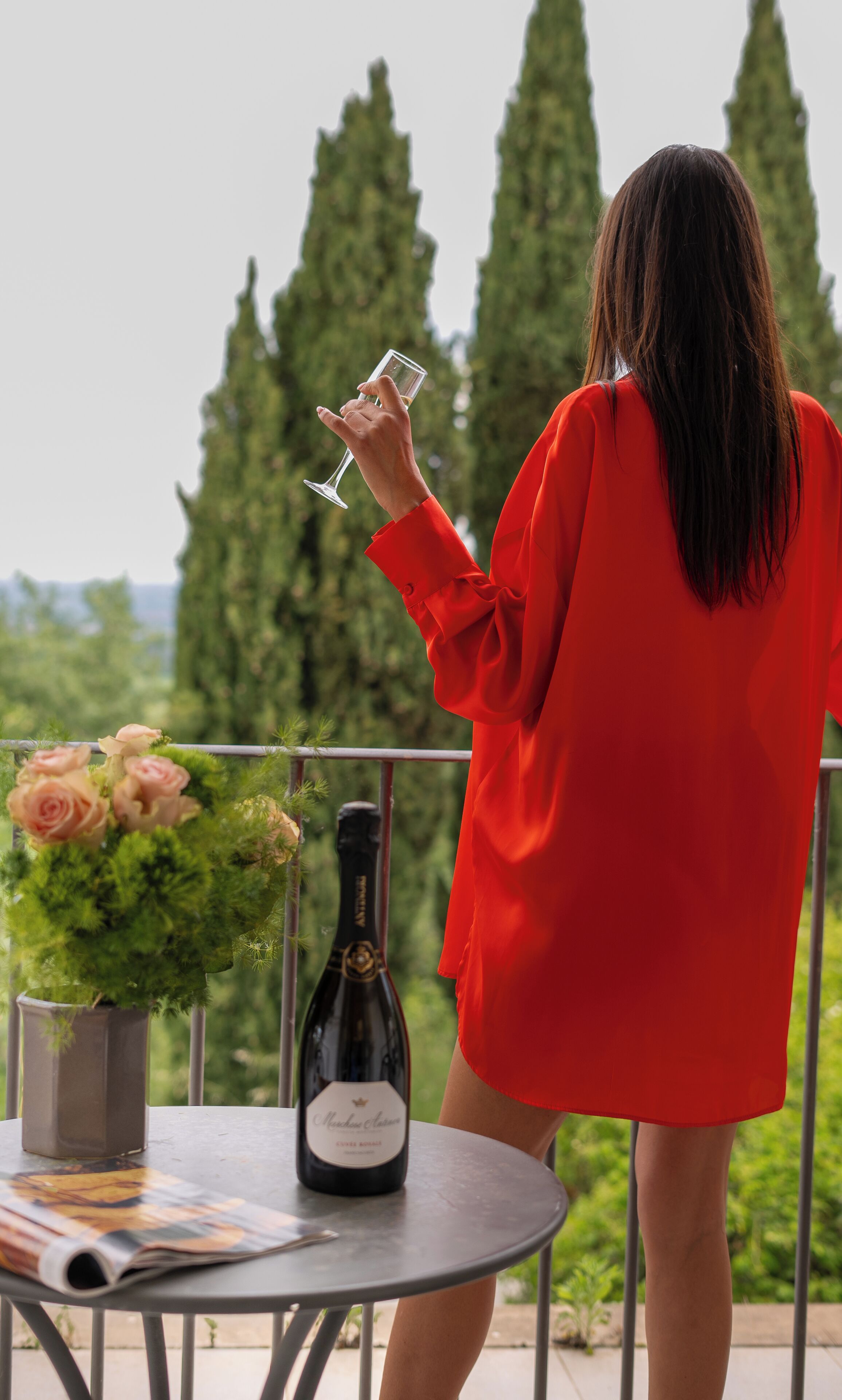 A person in a red dress standing at a table with a bottle of wine, a glass, and a vase of flowers.