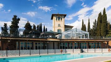 Una piscina con zona acristalada, una torre y altos árboles al fondo.