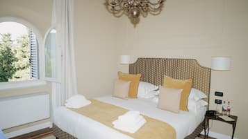A hotel room with a large bed, a chandelier, a bedside table with a lamp, and a window with curtains.