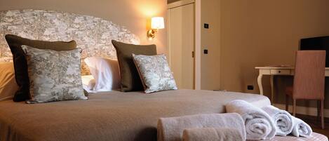 A neatly made bed with multiple pillows, a bedside table, a chair, and a desk with a lamp.