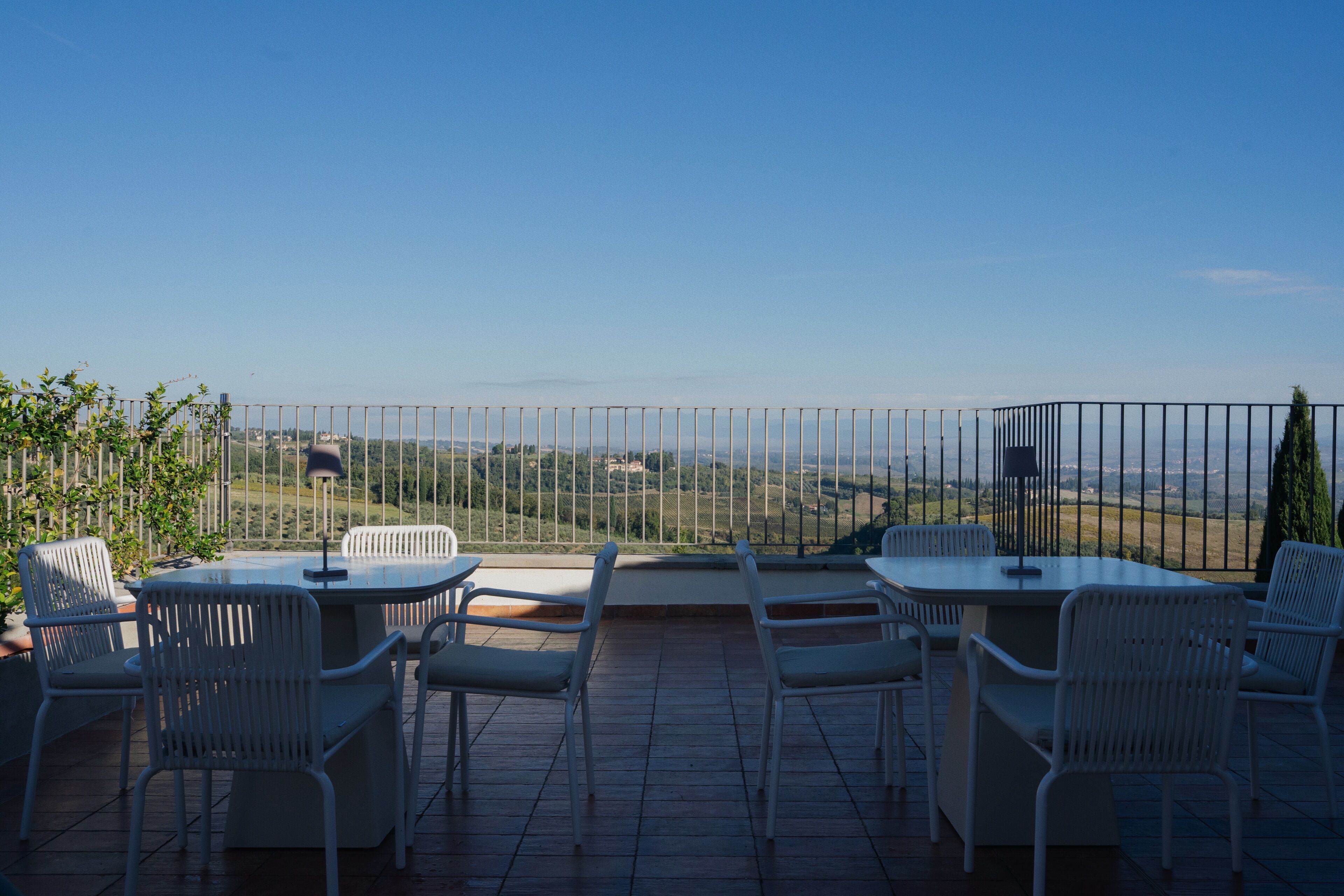 Um terraço com cadeiras e mesas brancas, uma cerca de metal e vista para o campo.