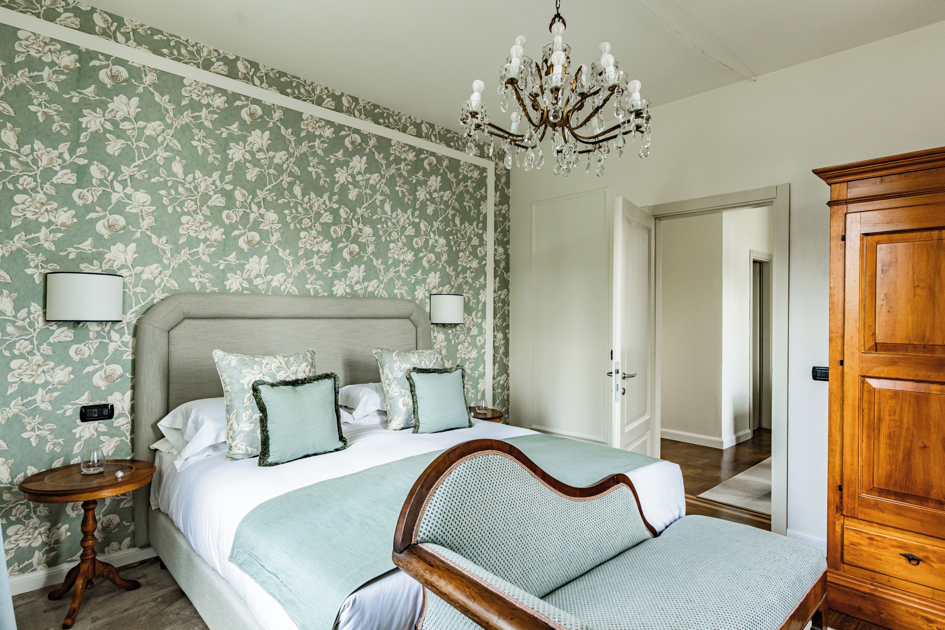 Un dormitorio con una cama grande, papel tapiz floral, un candelabro y un armario de madera.