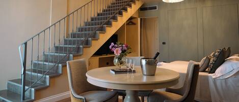 A hotel room with a round dining table, two chairs, a chandelier, a bed with pillows, and a staircase.